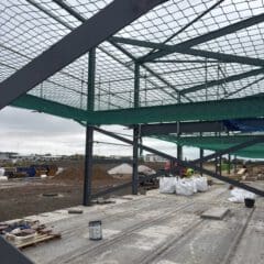 A construction site with a steel building framework, safety nets overhead, and construction materials scattered on the concrete floor. Workers in high-visibility clothing are present in the background. The sky is cloudy.