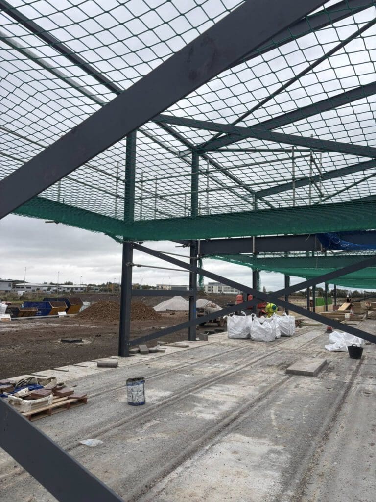 A construction site with a steel building framework, safety nets overhead, and construction materials scattered on the concrete floor. Workers in high-visibility clothing are present in the background. The sky is cloudy.