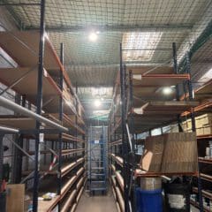 A warehouse aisle with tall metal shelves, some stacked with boxes and containers, others mostly empty. A blue ladder stands at the end of the aisle under a ceiling with protective netting and fluorescent lighting.