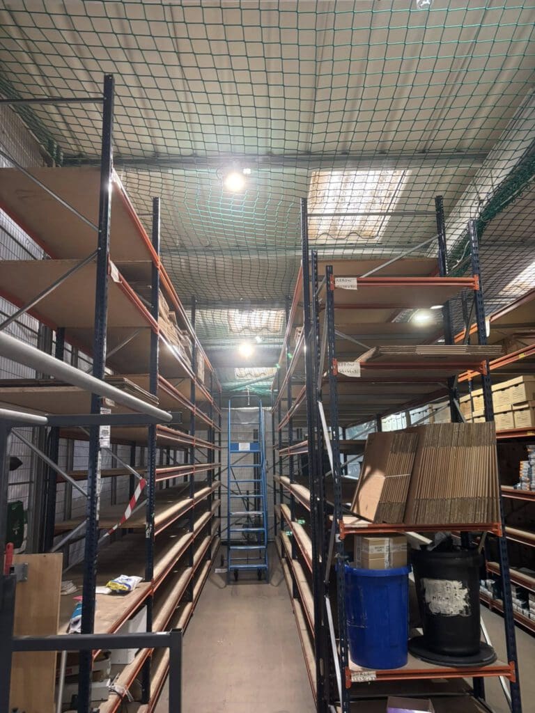 A warehouse aisle with tall metal shelves, some stacked with boxes and containers, others mostly empty. A blue ladder stands at the end of the aisle under a ceiling with protective netting and fluorescent lighting.