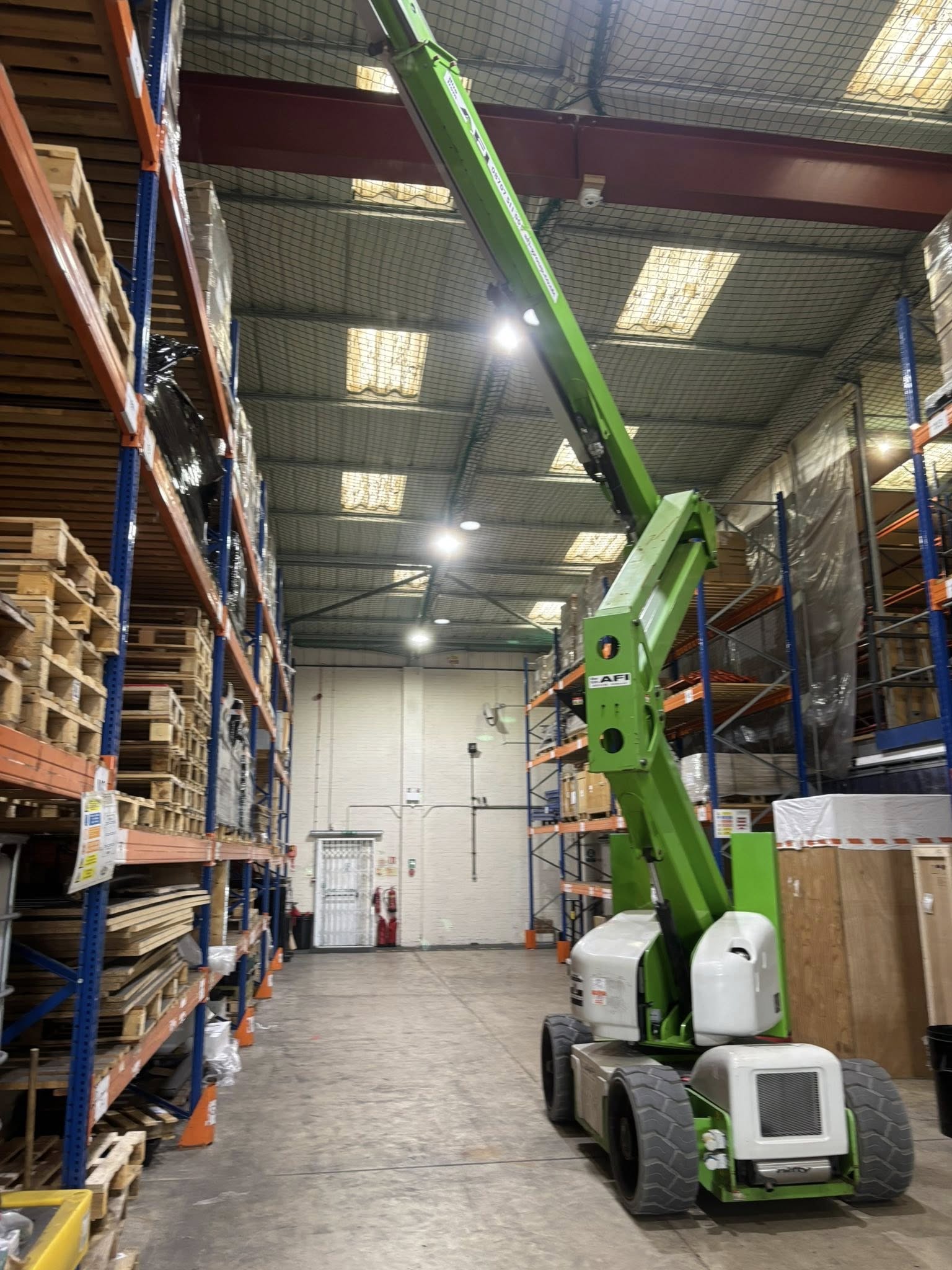 A green boom lift extends upward in a warehouse aisle surrounded by tall shelving units stacked with wooden pallets and boxes. The warehouse has high ceilings and fluorescent lighting.