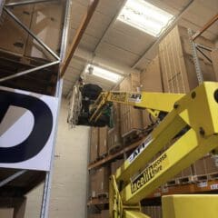 A yellow Facelift access lift is raised in a warehouse, reaching high shelves stacked with large boxes. The lift is positioned near the top shelf, and some equipment and cables are visible on the platform.