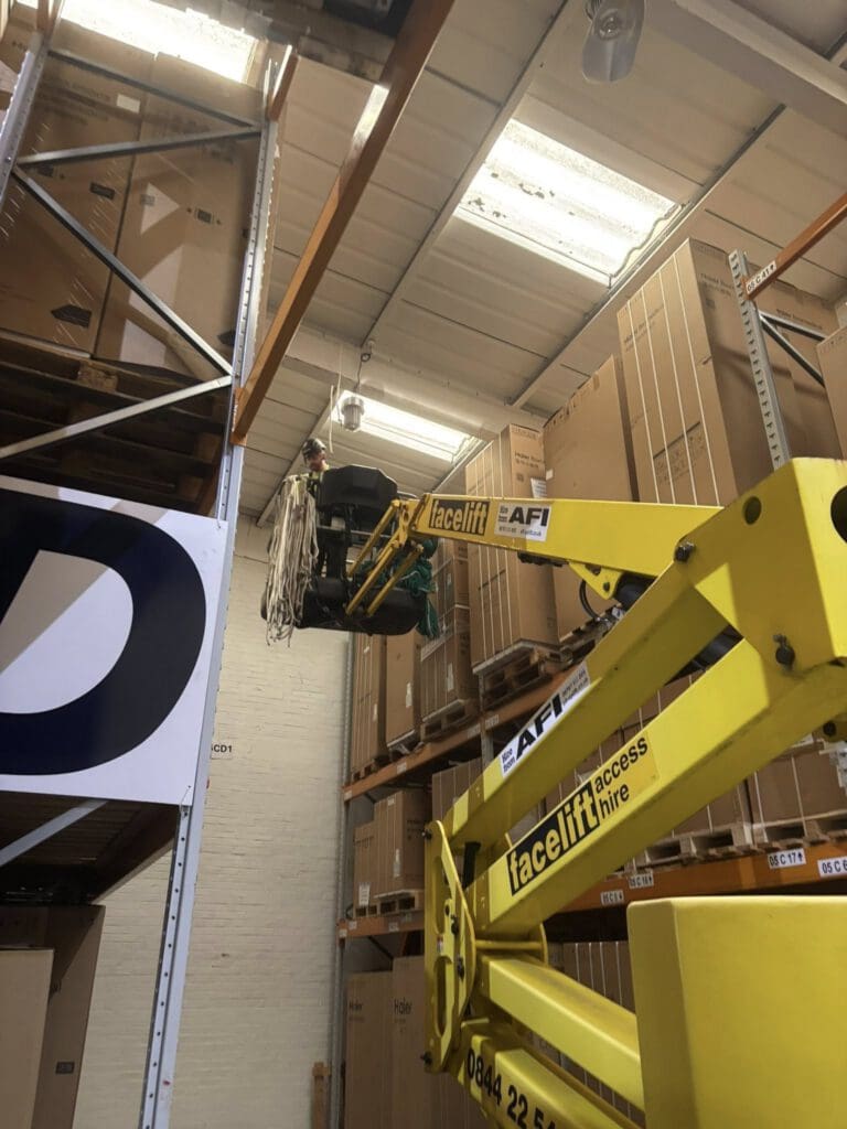 A yellow Facelift access lift is raised in a warehouse, reaching high shelves stacked with large boxes. The lift is positioned near the top shelf, and some equipment and cables are visible on the platform.