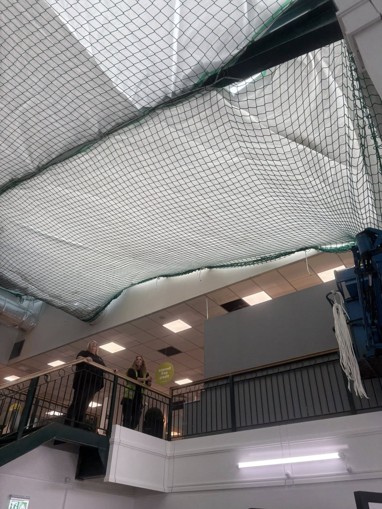 A large safety net is suspended from the ceiling above a staircase inside a building. Two people stand on the upper level near the railing, looking up at the net. Ceiling tiles are visible above the net.