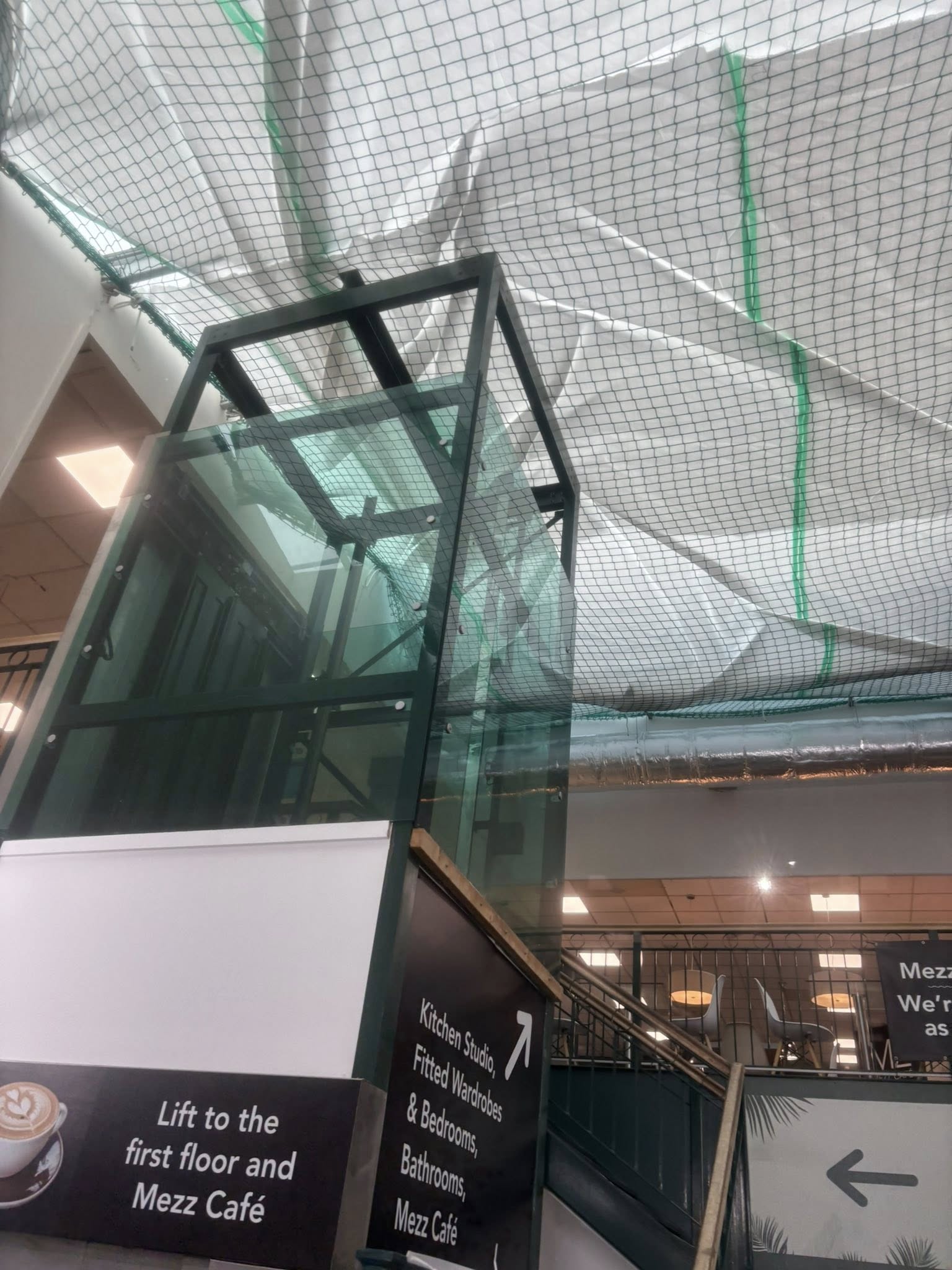 A glass lift next to a sign pointing to the first floor and Mezz Café, with protective netting and plastic sheeting overhead in a building under renovation.