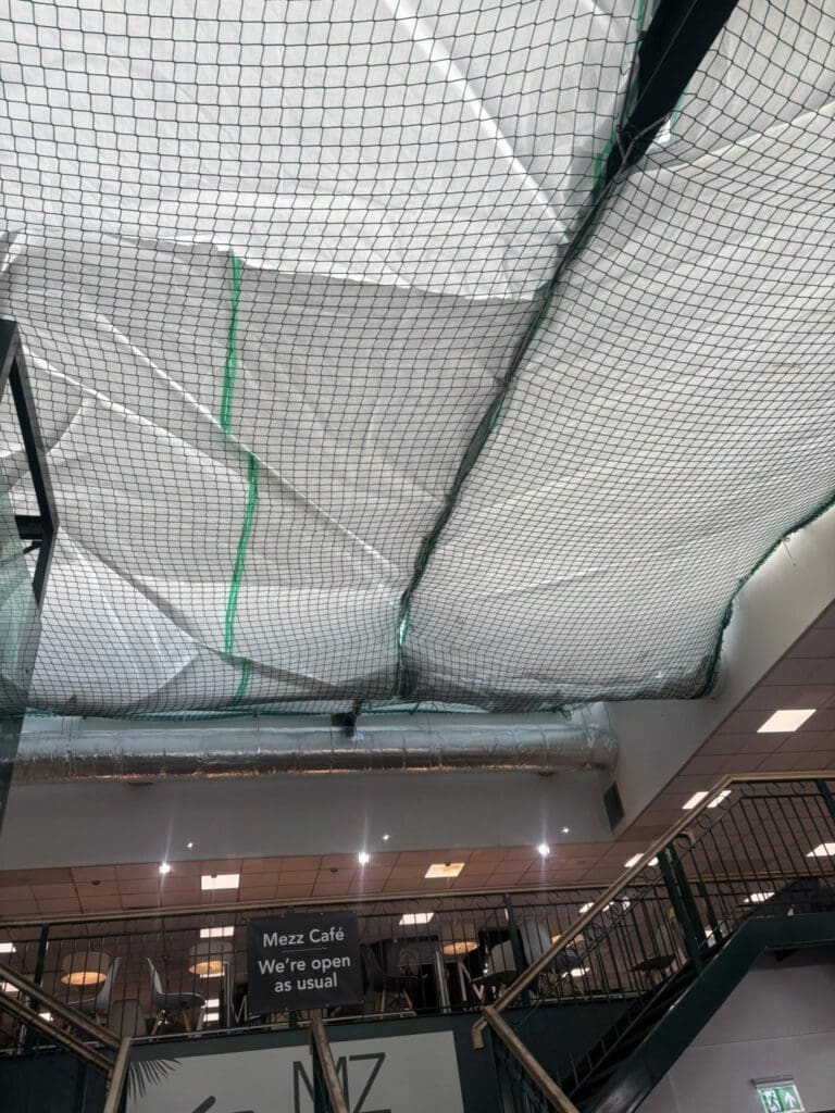 A ceiling covered with netting and white sheeting, likely for maintenance or construction. Below, a sign reads "Mezz Café We're open as usual," and stairs lead up to the café area.