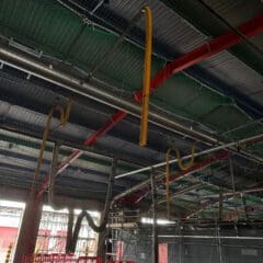 Large industrial space with exposed pipes and ducts hanging from the ceiling, some yellow and curved. Red metal beams and scaffolding are visible, along with red barriers on the floor. The area appears under construction or renovation.