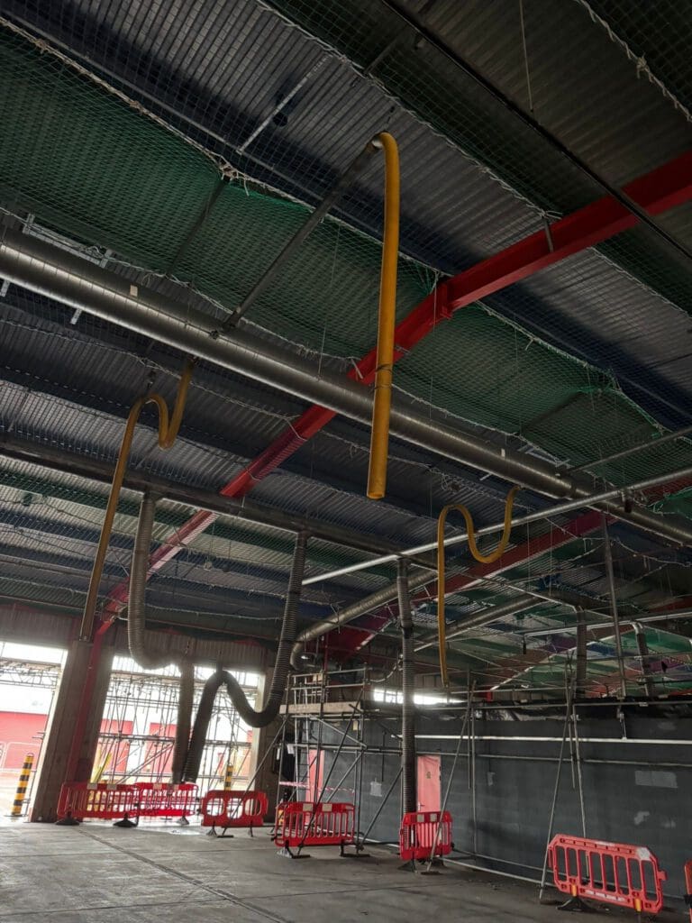 Large industrial space with exposed pipes and ducts hanging from the ceiling, some yellow and curved. Red metal beams and scaffolding are visible, along with red barriers on the floor. The area appears under construction or renovation.