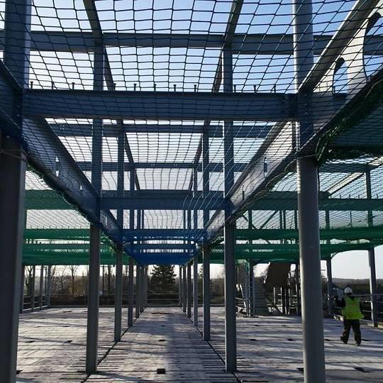 A construction site shows a steel framework of a building with safety nets suspended between beams. Two workers in safety gear are visible on the right, and the sky is clear in the background.