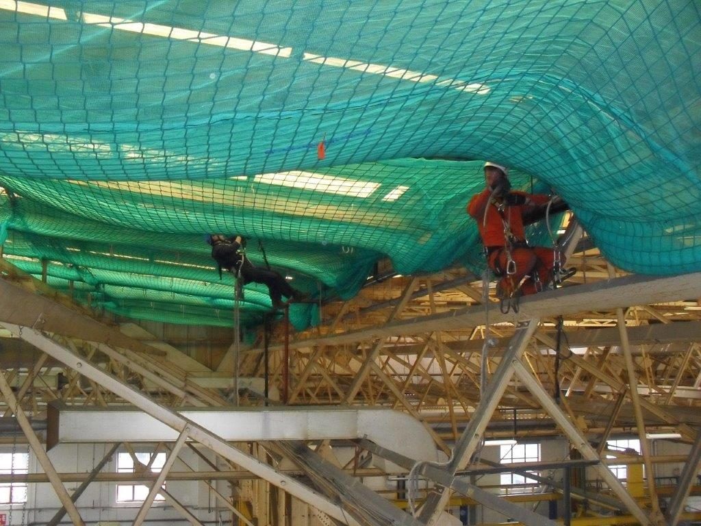 Two workers in safety harnesses are suspended above beams inside a large industrial building, working on or inspecting green netting installed near the ceiling for safety or construction purposes.
