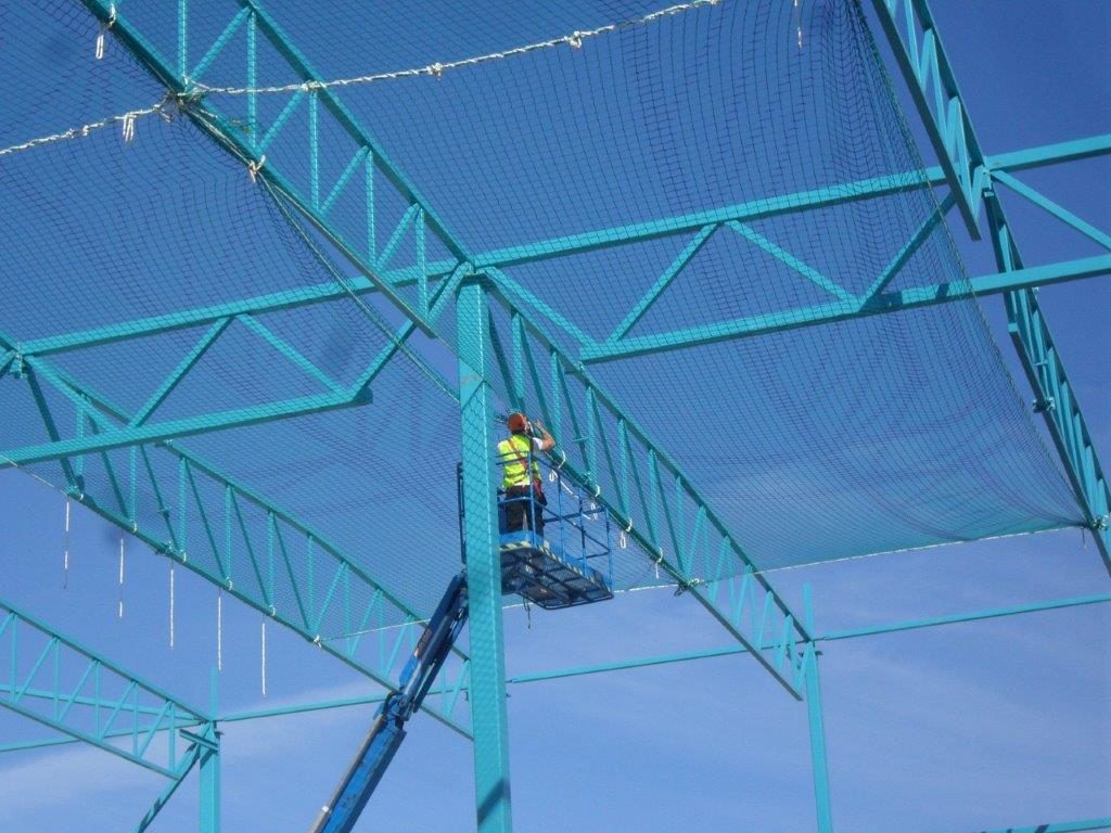 A construction worker in a safety vest stands on a raised platform, installing or inspecting netting on a blue steel building framework under a clear sky.