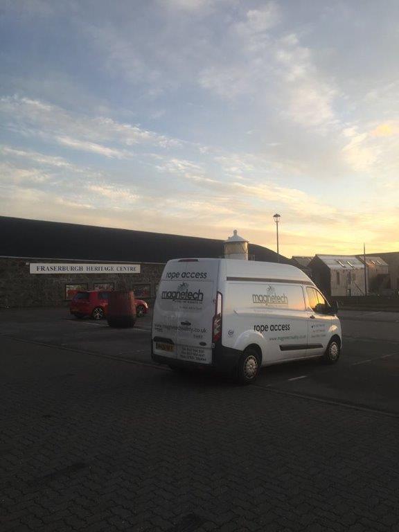 A white van with "magpienetec" and "rope access" branding is parked in a lot near the Fraserburgh Heritage Centre, with old stone buildings and a cloudy sky at sunset in the background.