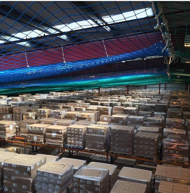 Large warehouse filled with stacks of cardboard boxes on pallets, organized in rows. Overhead, a blue and green safety net is suspended from the ceiling, illuminated by skylights and ceiling lights.