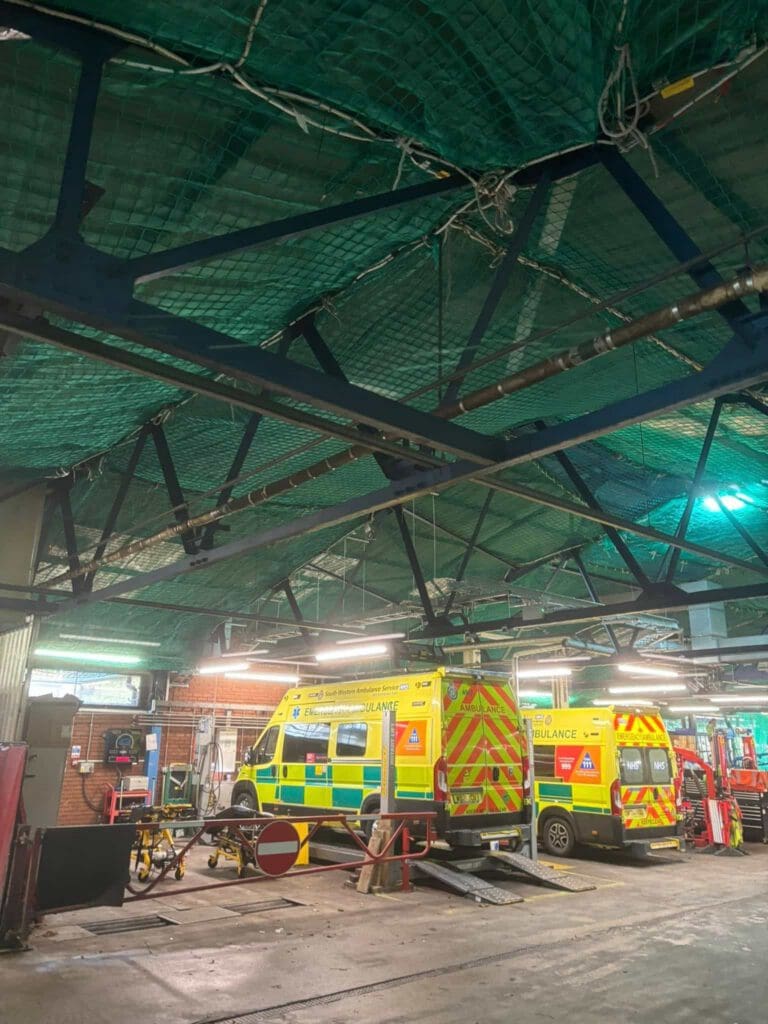 Two ambulances with yellow and green markings are parked inside a garage with exposed beams and green netting on the ceiling. The area is lit by fluorescent lights, and equipment is visible in the background.