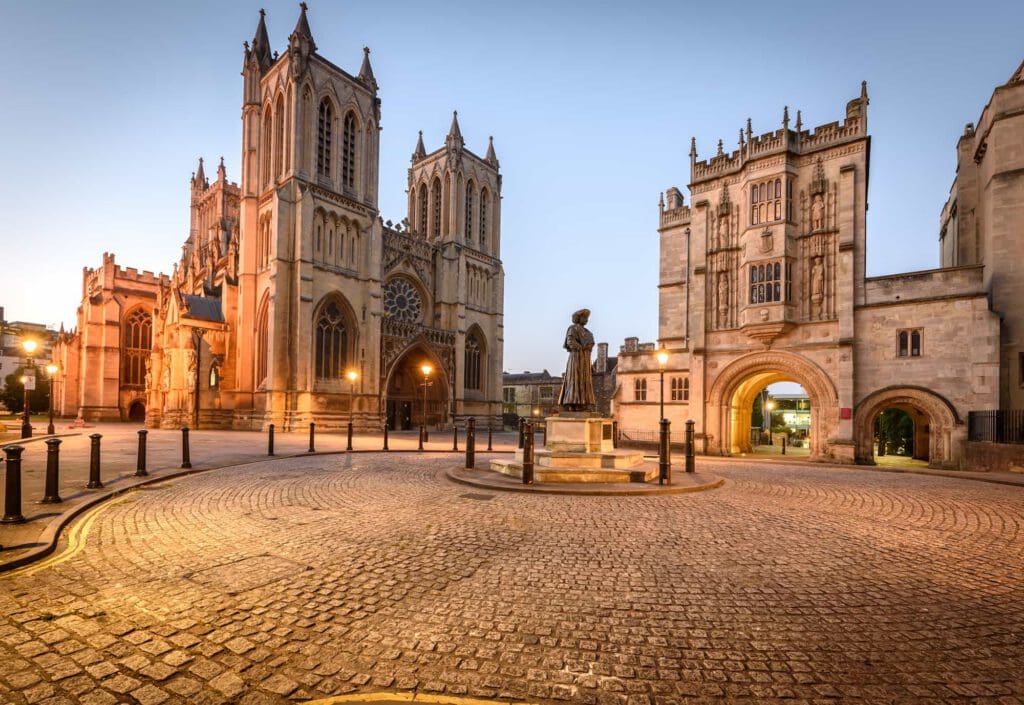 A cobblestone square at dusk features a statue in the center, surrounded by historic stone buildings with arched windows, including a large cathedral and a grand arched gateway.