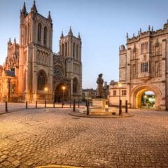 A cobblestone square at dusk features a statue in the center, surrounded by historic stone buildings with arched windows, including a large cathedral and a grand arched gateway.