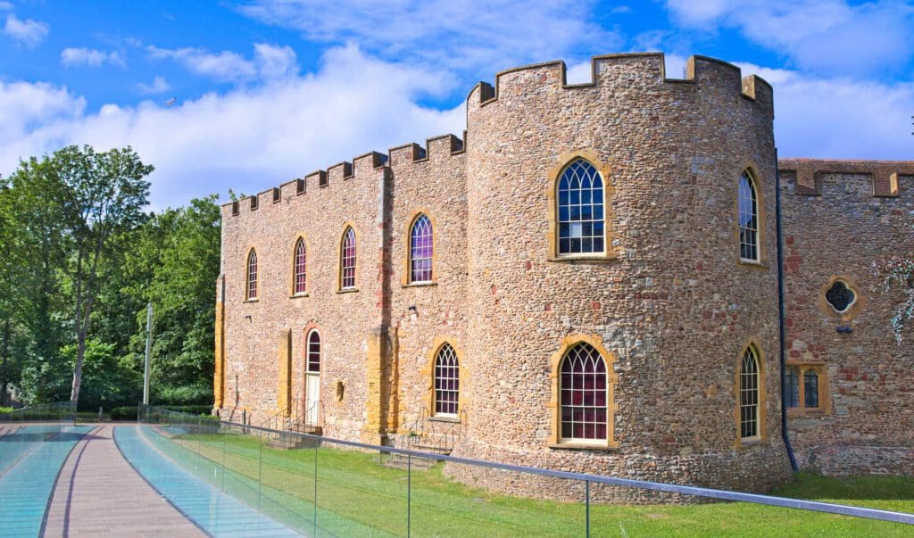 A historic stone castle with arched windows and crenellated towers stands beside a modern glass walkway, surrounded by green grass and trees under a bright blue sky with scattered clouds.