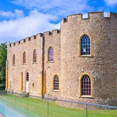 A historic stone castle with arched windows and crenellated towers stands beside a modern glass walkway, surrounded by green grass and trees under a bright blue sky with scattered clouds.