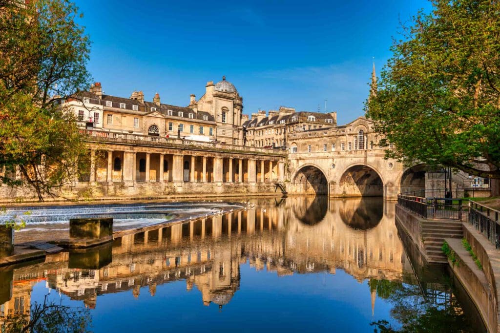 Historic stone buildings and the Pulteney Bridge reflect in the calm water of the River Avon in Bath, England, under a clear blue sky with lush green trees framing the scene.