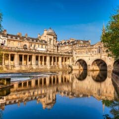 Historic stone buildings and the Pulteney Bridge reflect in the calm water of the River Avon in Bath, England, under a clear blue sky with lush green trees framing the scene.