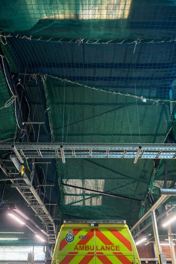 An ambulance with high-visibility markings is parked under a ceiling covered with green netting and metal framework, indicating ongoing construction or renovation work above.