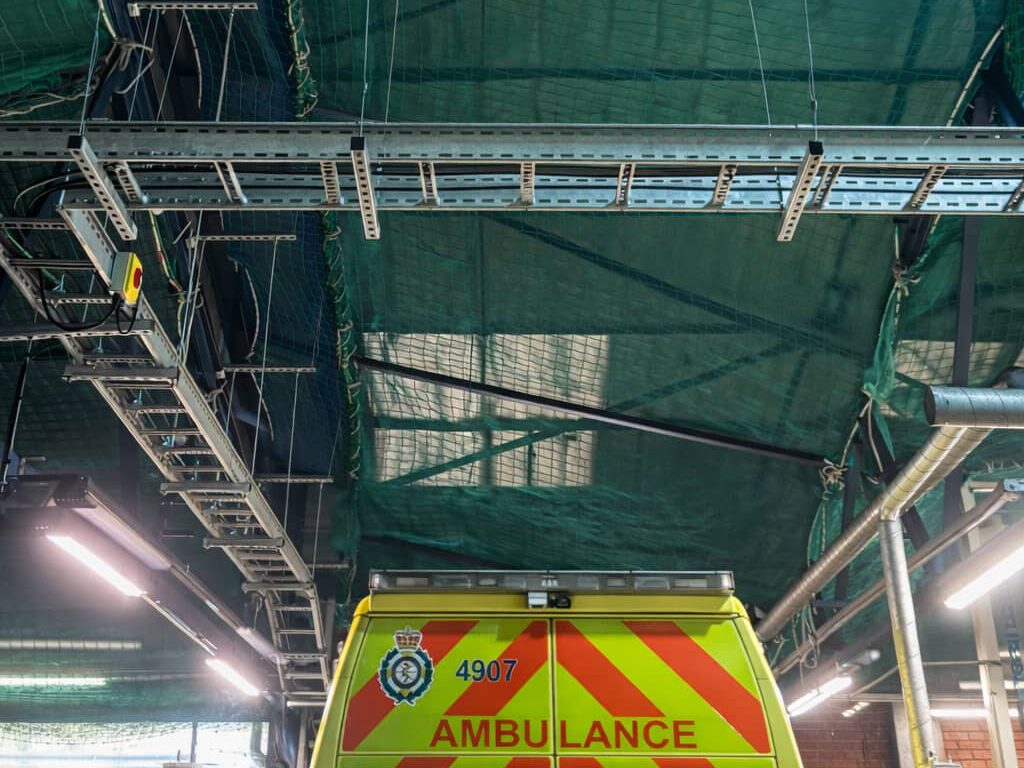 The rear of an ambulance with red and yellow chevrons is parked inside a building with green netting and exposed metal beams overhead.