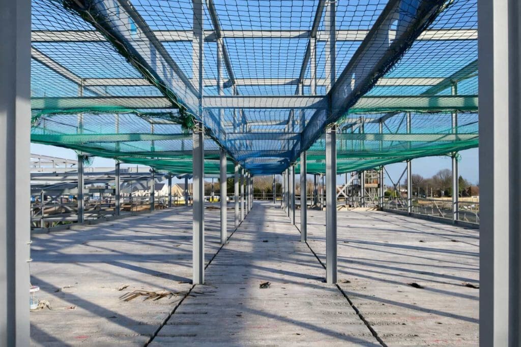 A construction site shows a concrete floor with metal beams and vertical columns, covered by green and blue safety nets overhead. The area is open and sunlit with building materials scattered on the ground.