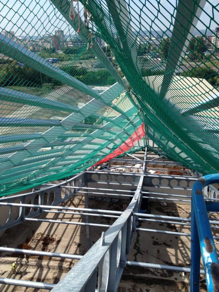 View from a high structure showing steel beams, green safety nets overhead, and a partially constructed platform, with a cityscape and greenery visible in the background.