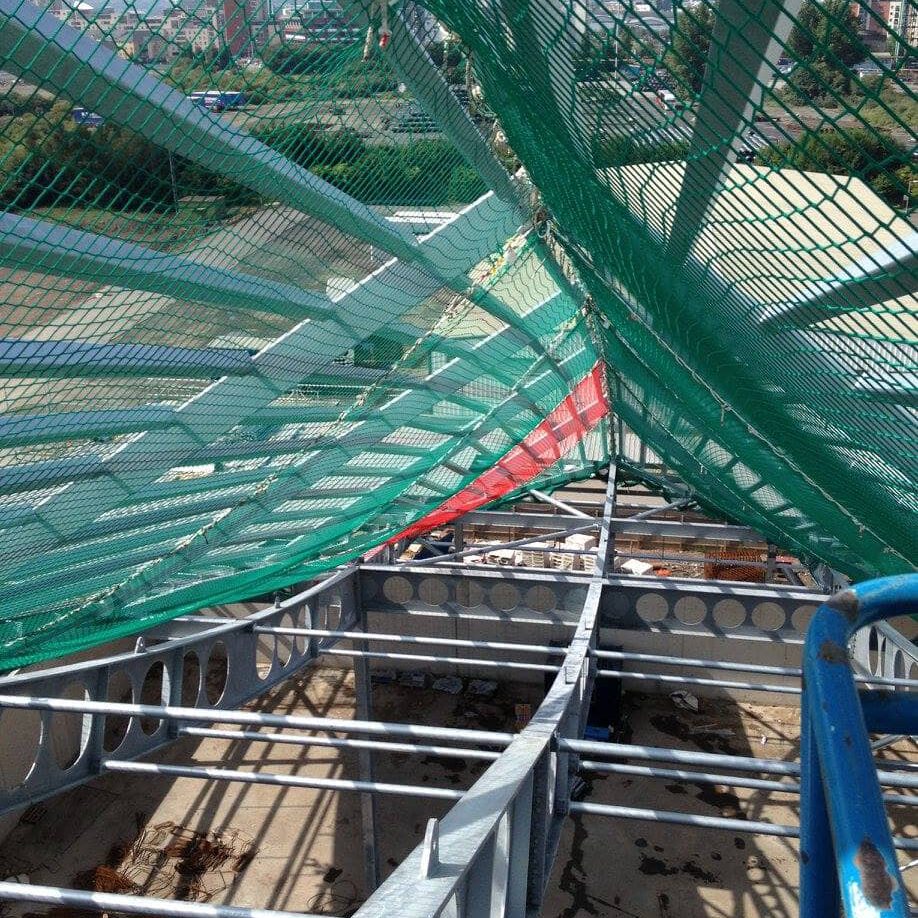View from above a construction site showing metal beams, a safety net stretched overhead, and some construction materials and vehicles below. The net provides protection for workers on the structure.