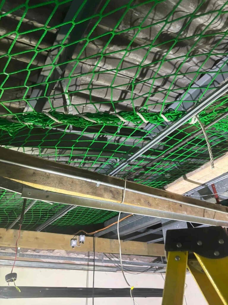 A green safety net is installed beneath a ceiling with exposed insulation, metal beams, and electrical wiring. A yellow ladder is positioned in the lower right corner of the image.