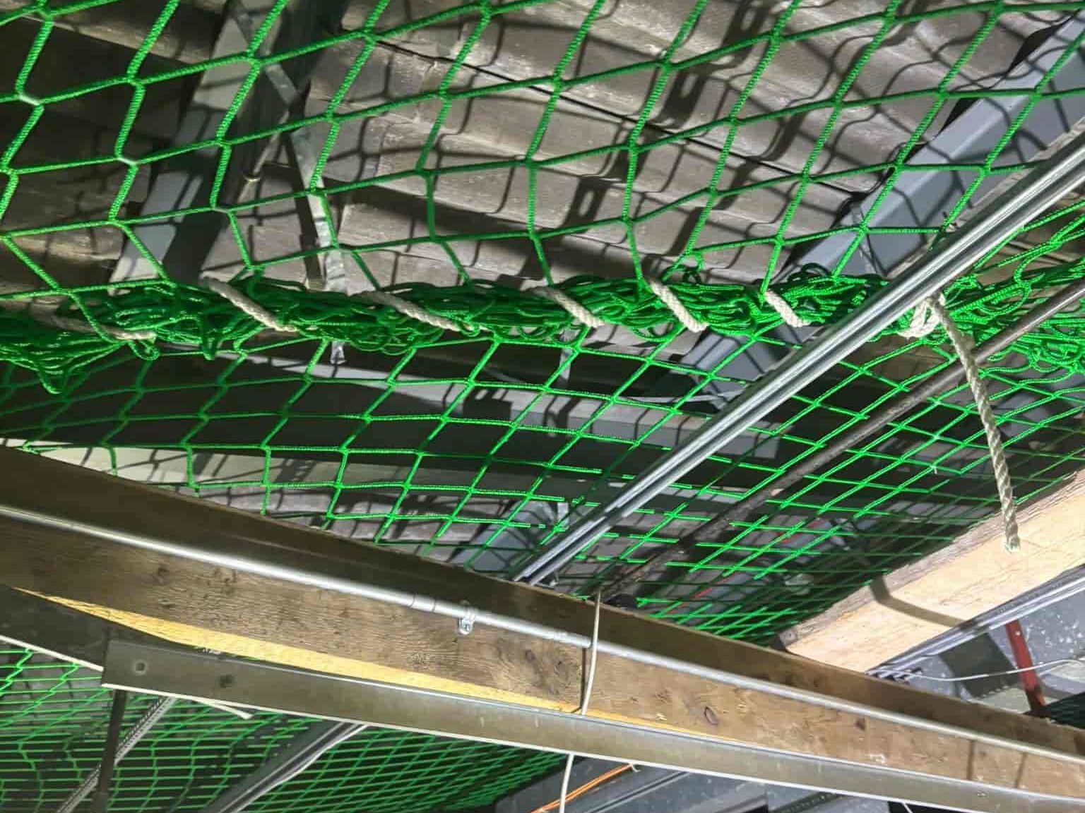 Green safety netting is suspended above a construction area, with metal pipes and wooden beams running horizontally underneath. The structure above the netting is partially visible.