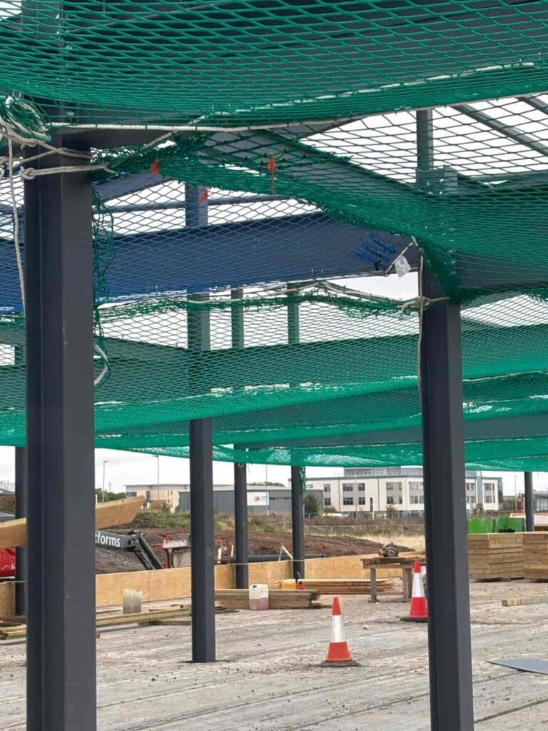 Construction site with green safety netting secured to steel beams, orange traffic cones, and building materials on the ground. Buildings and cloudy sky visible in the background.