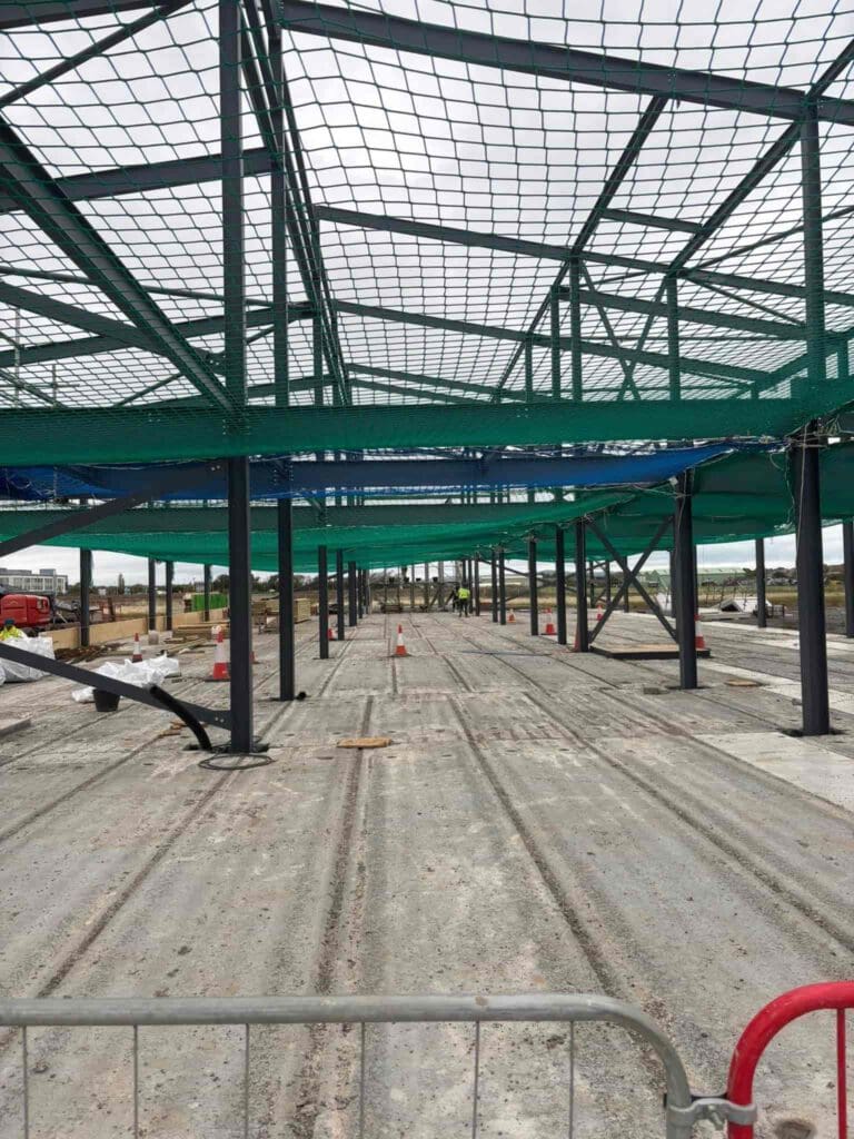 Steel framework of a building under construction, with green safety nets overhead and unfinished ground below. Construction materials and workers are visible in the background, and a metal barrier is in the foreground.