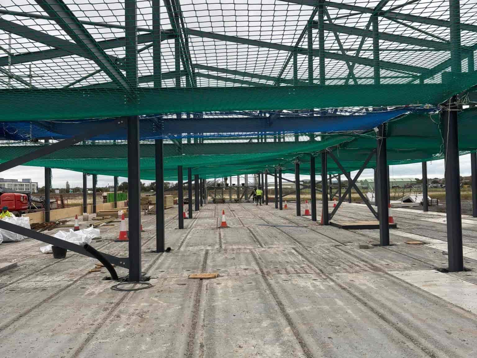 A construction site with steel framework, safety nets, and traffic cones on a concrete floor. Workers are visible in the distance, and the area is open to the outdoors with cloudy skies.