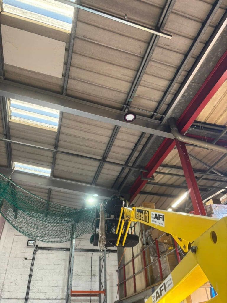 A yellow AFI nacelle lift extends towards the ceiling of an industrial warehouse with exposed beams, skylights, and a green safety net hanging near the roof.