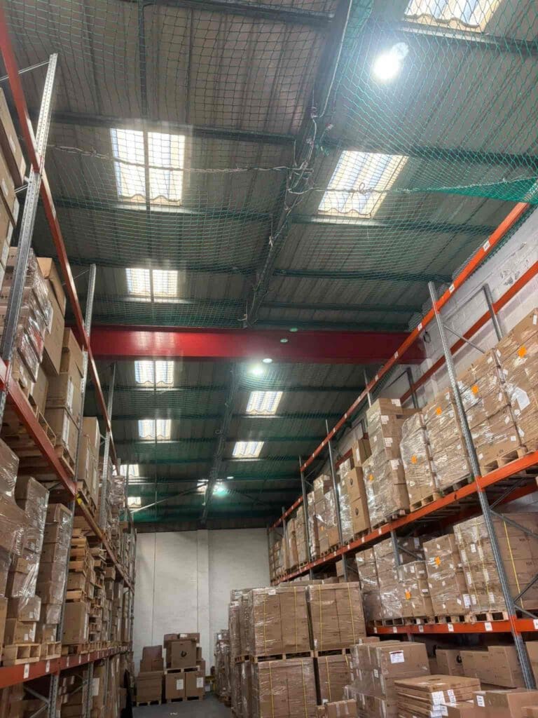A warehouse with high shelves stacked with cardboard boxes, fluorescent lights on the ceiling, and safety netting overhead. The perspective is from the floor, looking up at the shelves and ceiling.
