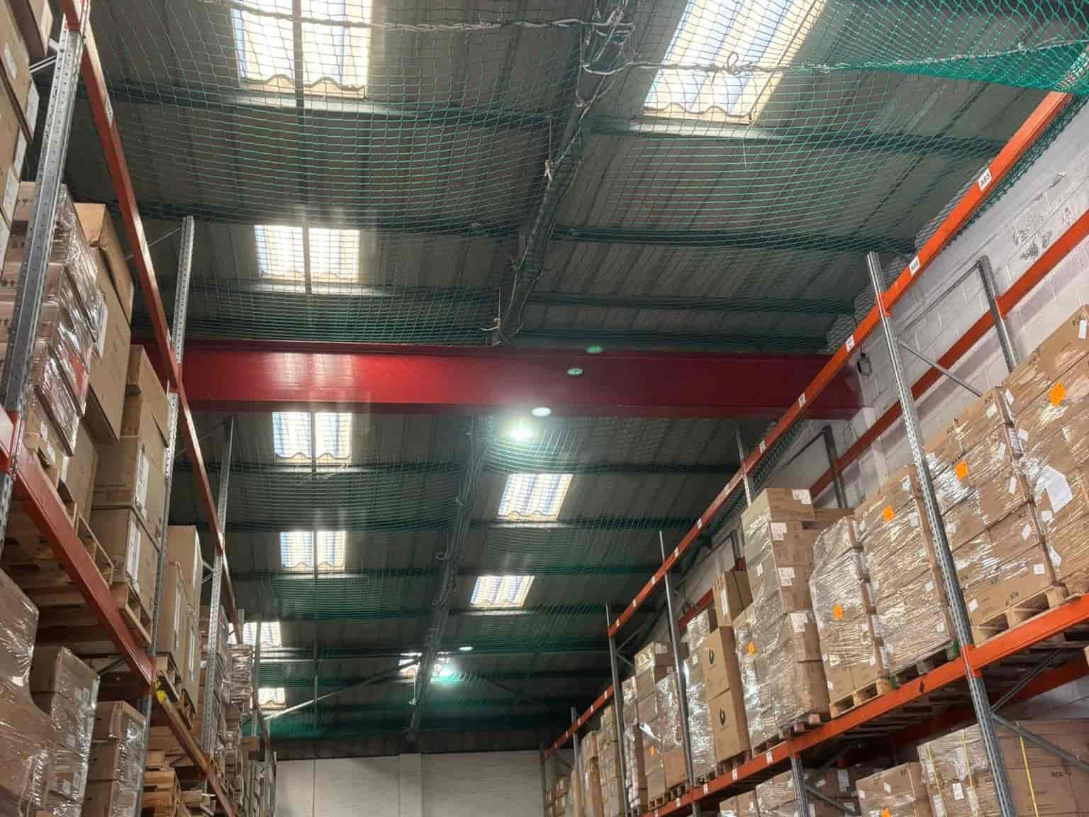 Warehouse interior with tall shelves stacked with cardboard boxes on both sides, overhead fluorescent lights, and a green safety net attached to the metal ceiling.