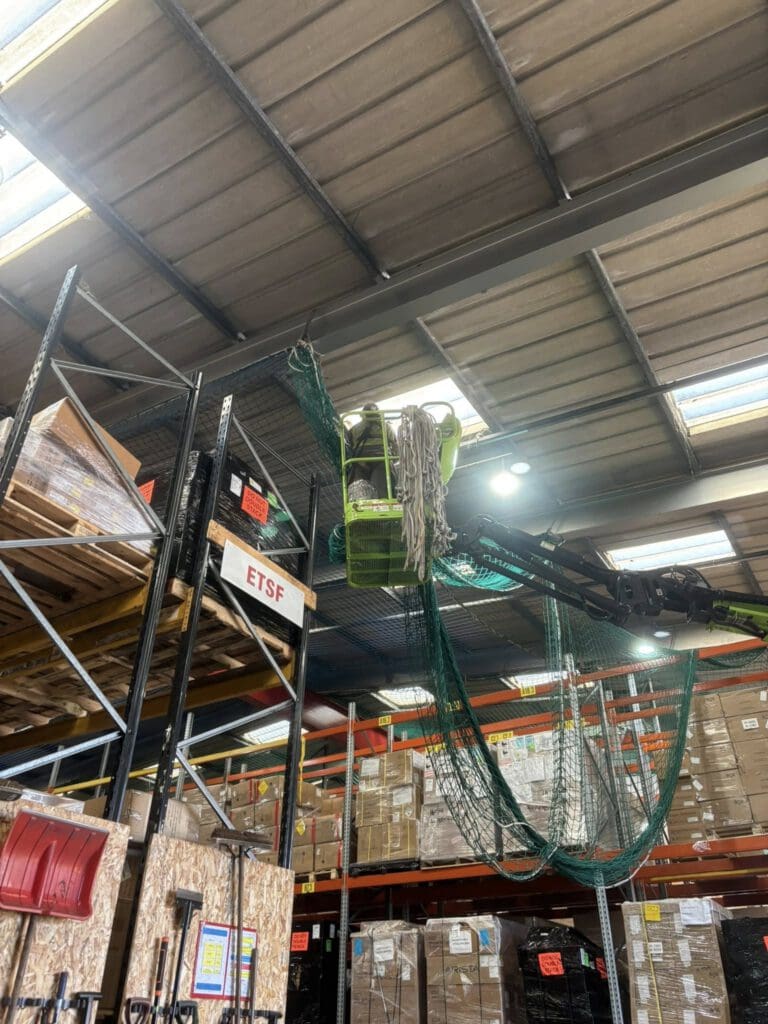 A worker in a bright vest operates a green lift to install or adjust a green safety net on high warehouse shelves, surrounded by boxes and industrial equipment under a metal roof with skylights.