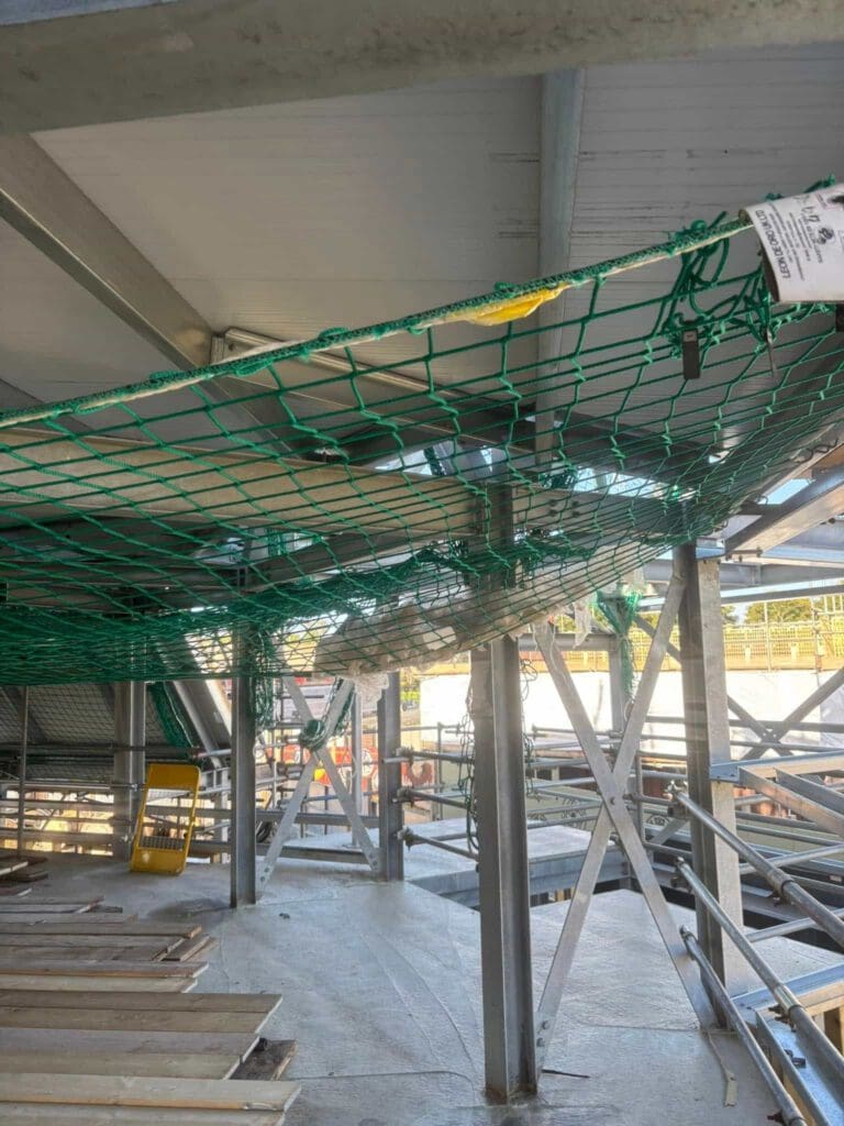 A green safety net is suspended under a metal structure at a construction site, partially sagging and holding debris. Sunlight filters in, and construction materials and equipment are scattered on the floor below.