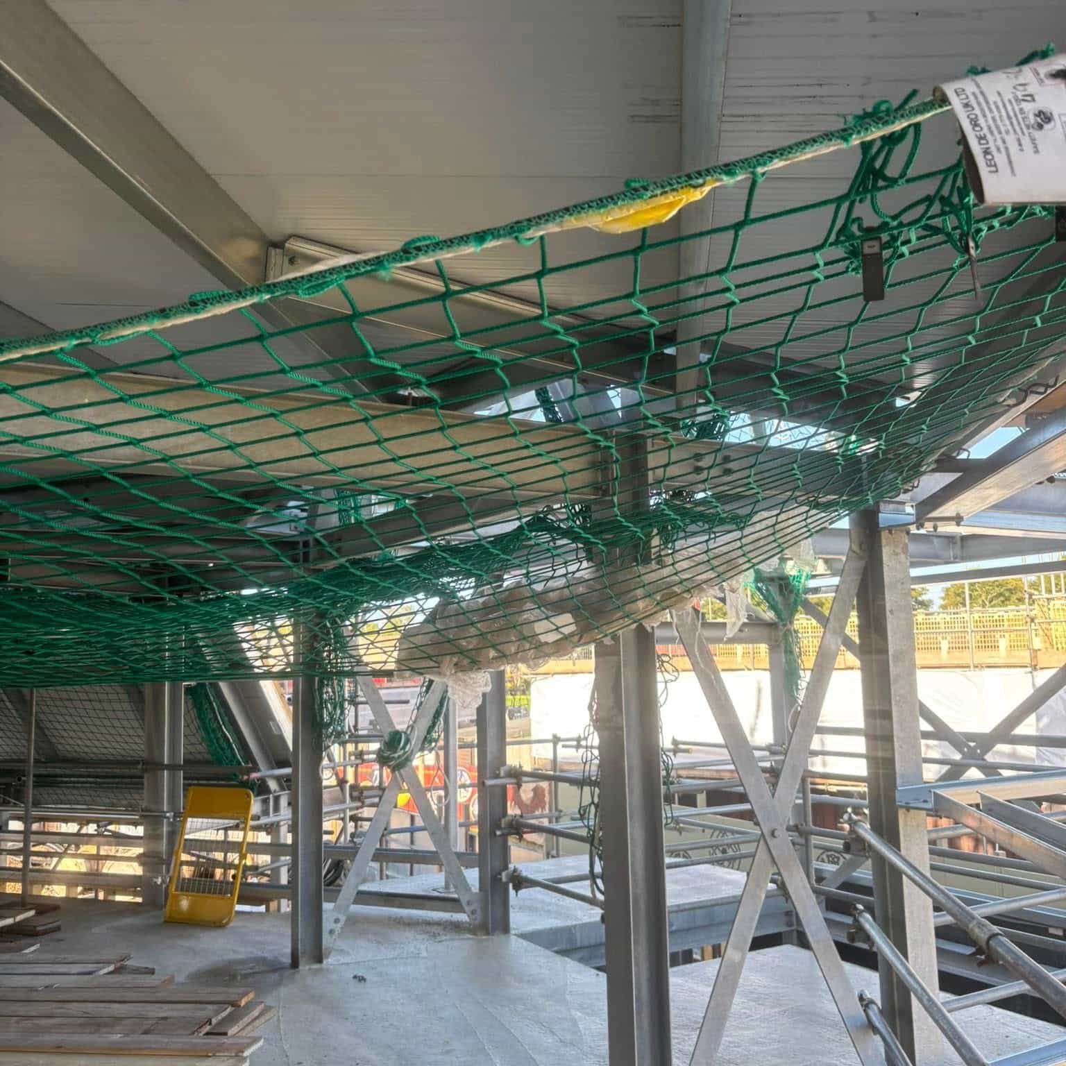 A green safety net is suspended under a metal roof structure at a construction site, catching a large fallen ceiling panel. Scaffolding and construction materials are visible in the background.