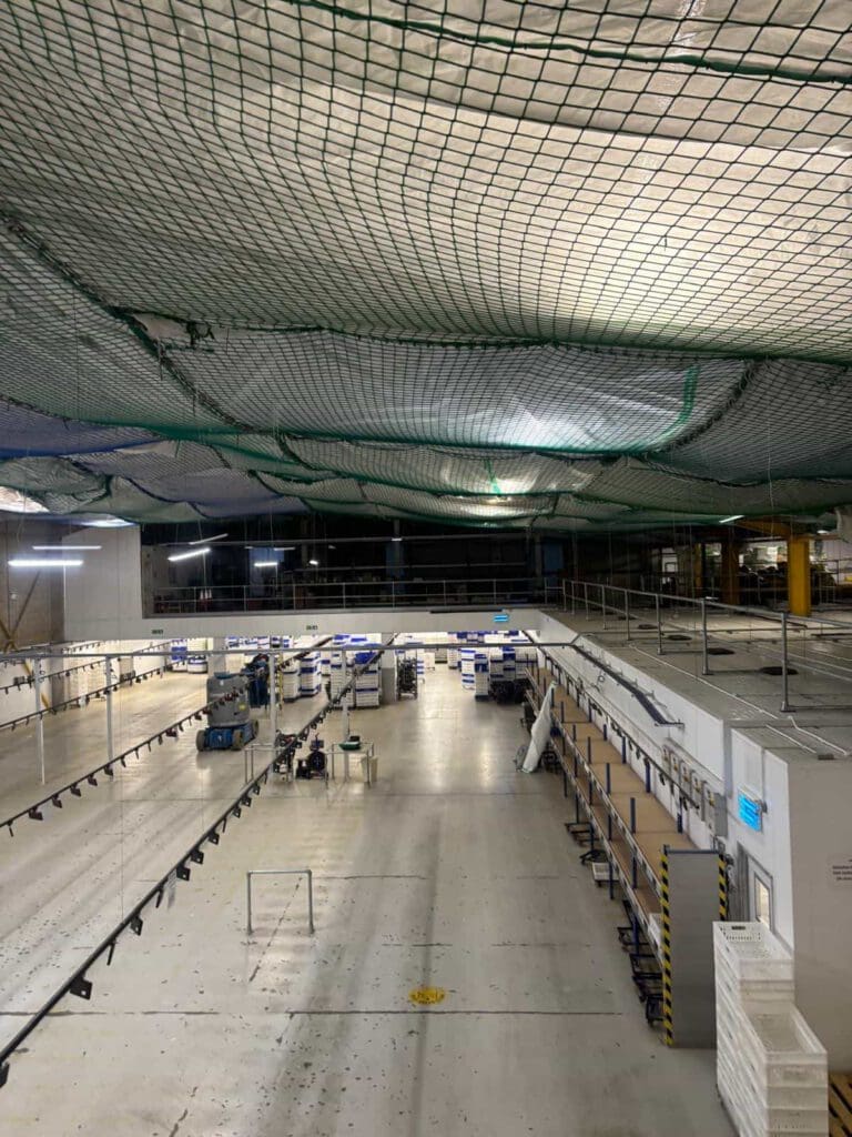 Large, empty warehouse interior with high ceilings and safety nets overhead. Rows of metal shelving units and workstations are visible below, with some scattered equipment and bins. The space is brightly lit and spacious.