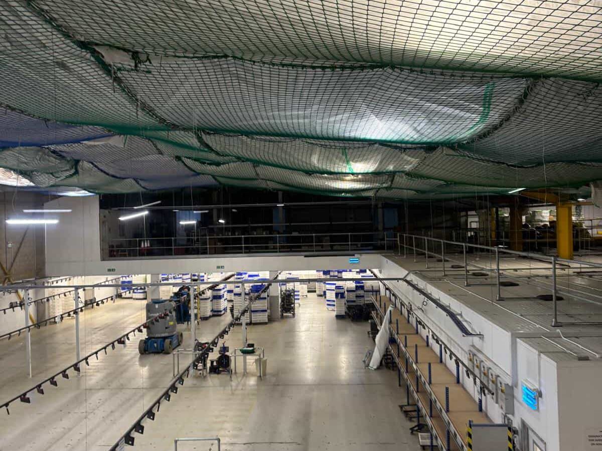 A large industrial warehouse with high ceilings, safety nets overhead, vacant workstations, and scattered equipment and boxes across the spacious floor. Bright ceiling lights illuminate the area.