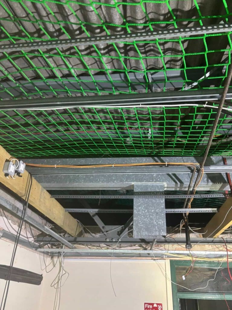 Exposed ceiling with metal beams, electrical wiring, and cables. Green safety netting is installed above the beams, and some pipes and conduits are visible. The ceiling tiles are missing, revealing the infrastructure above.