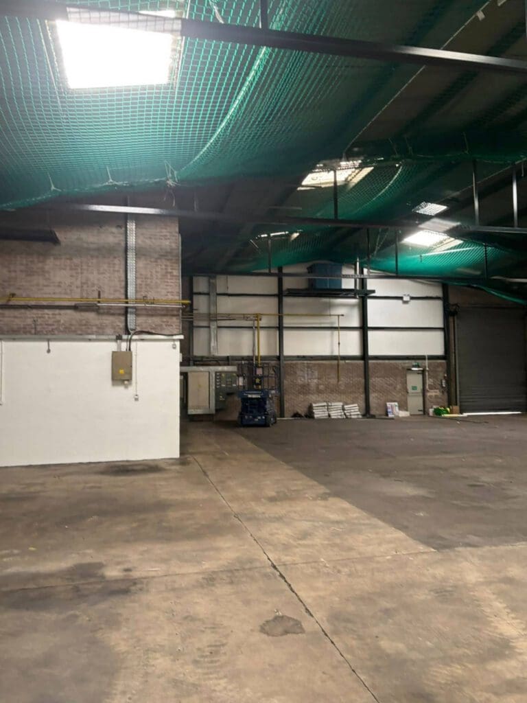 Interior of an empty industrial warehouse with concrete floors, brick walls, overhead lighting, green safety nets on the ceiling, and some stacked materials and equipment near the back wall.