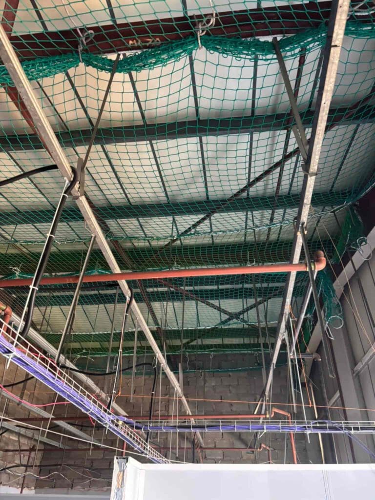 Green safety nets are installed across the ceiling of an industrial building under construction, with metal beams, wires, and pipes visible overhead.