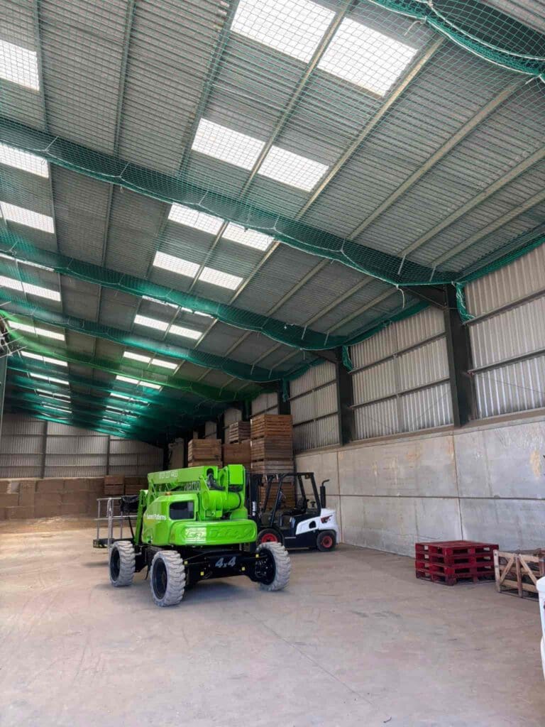 A spacious warehouse with high ceilings, stacks of wooden pallets and boxes along the back wall, a green lift vehicle, and a forklift parked on the concrete floor under skylights and green safety netting.