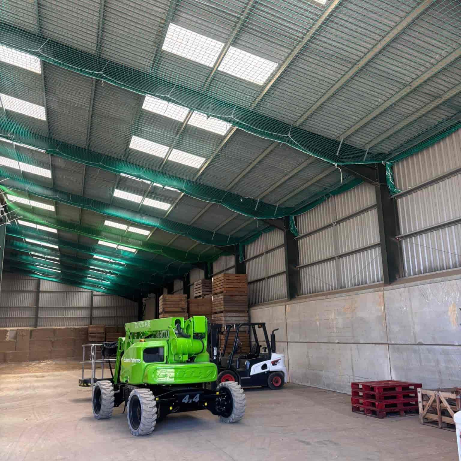 A large warehouse with high ceilings, skylights, and green netting overhead. Inside are a green telescopic boom lift, a black and white forklift, stacked pallets, and bales of material along the walls.