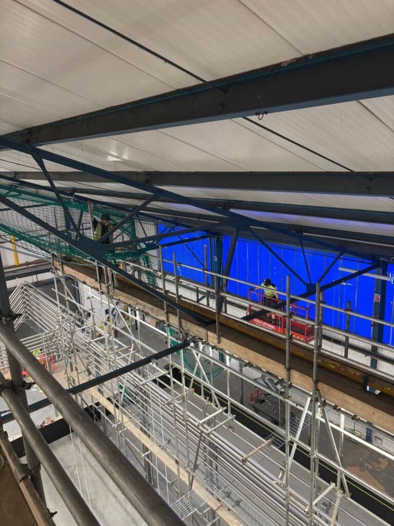 Construction site interior with scaffolding and workers wearing safety gear elevated near metal beams under a high ceiling, with blue partition walls in the background.