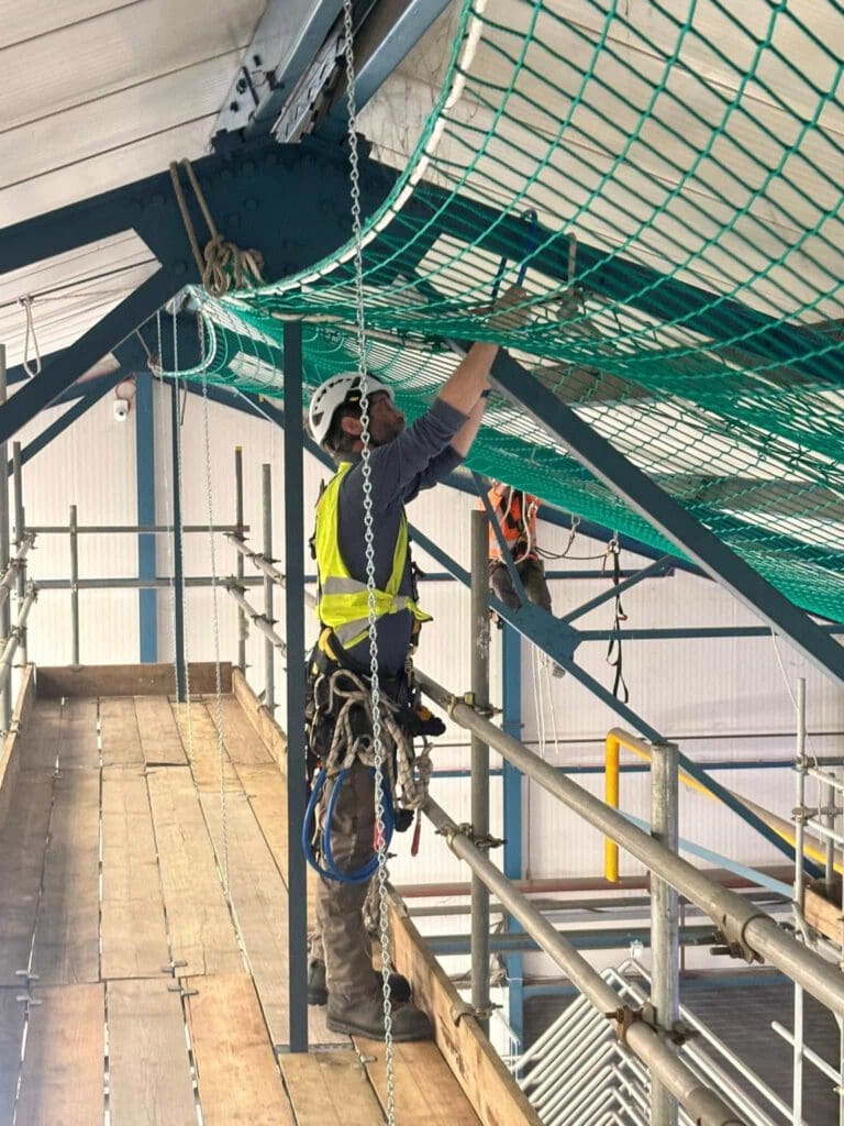 A construction worker in a safety harness and helmet secures green netting to a metal beam while standing on scaffolding inside a building. Another worker is visible in the background, also working with the netting.