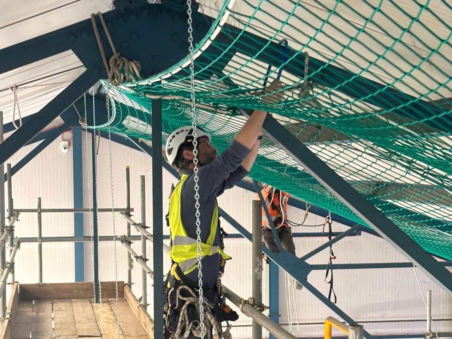 A worker wearing a helmet, safety harness, and high-visibility vest secures a green safety net to a metal beam inside an industrial building. Another worker is visible in the background on the scaffolding.